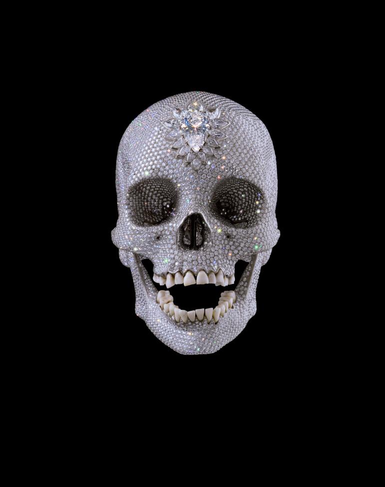 A platinum cast of a human skull covered entirely in diamonds, with a large pear-shaped diamond on the forehead and real teeth, against black.